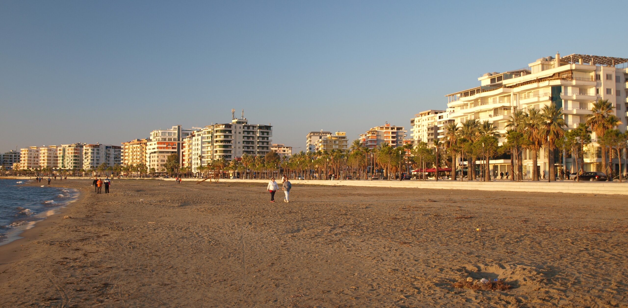 Everyday coastal city scene in Albania with apartment buildings, pedestrians, and the Adriatic or Ionian water nearby.