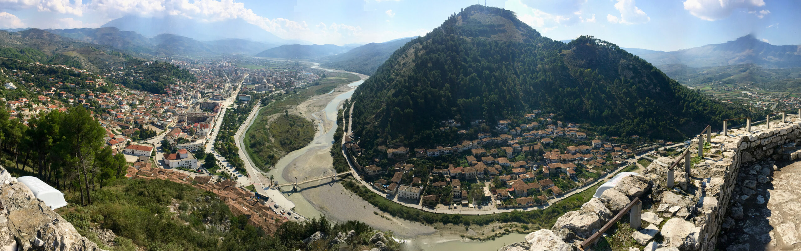 Wide daylight panorama over Berat with historic hillside homes and the river valley below.