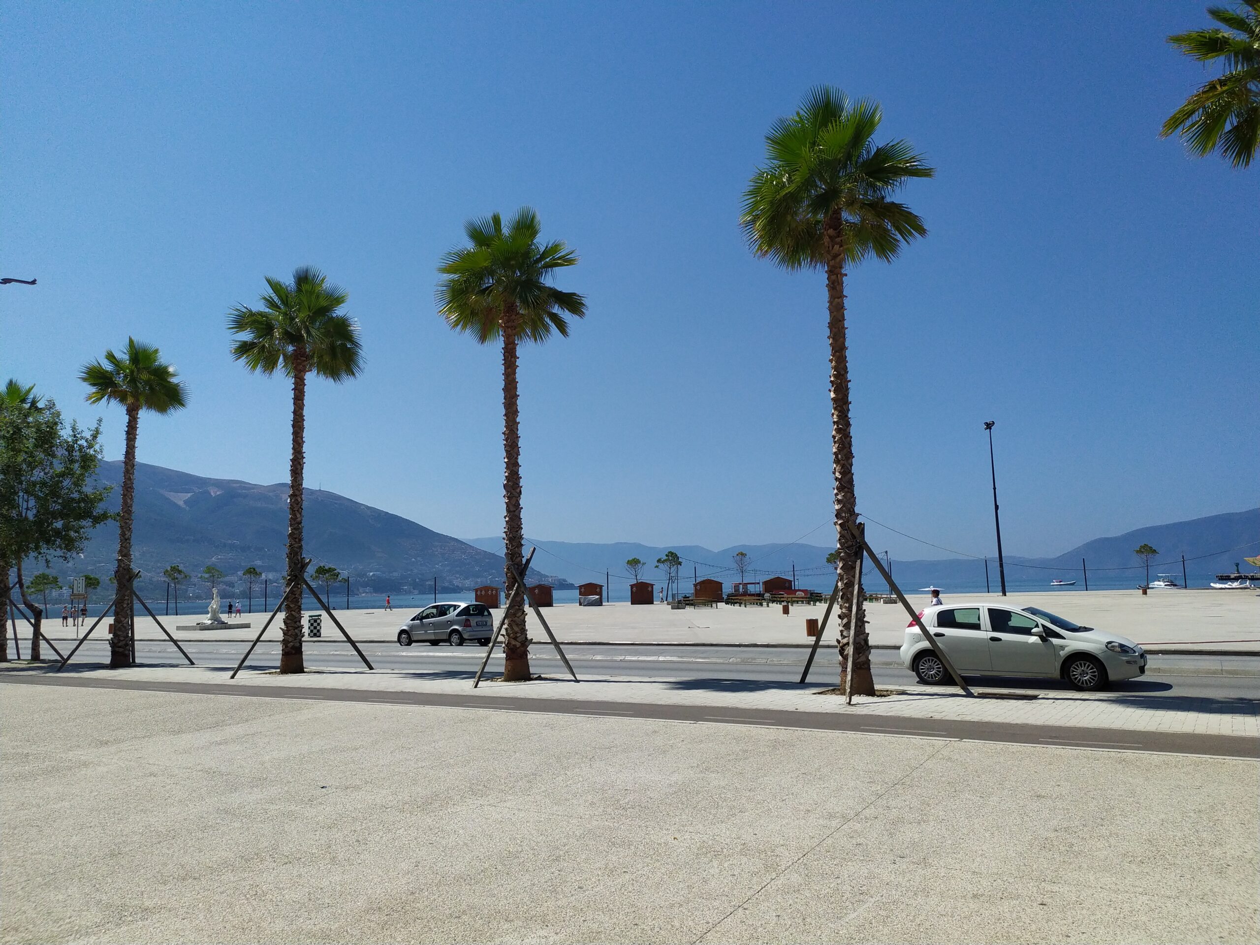 Palm-lined Lungomare waterfront in Vlorë with sea and mountains visible in daylight.