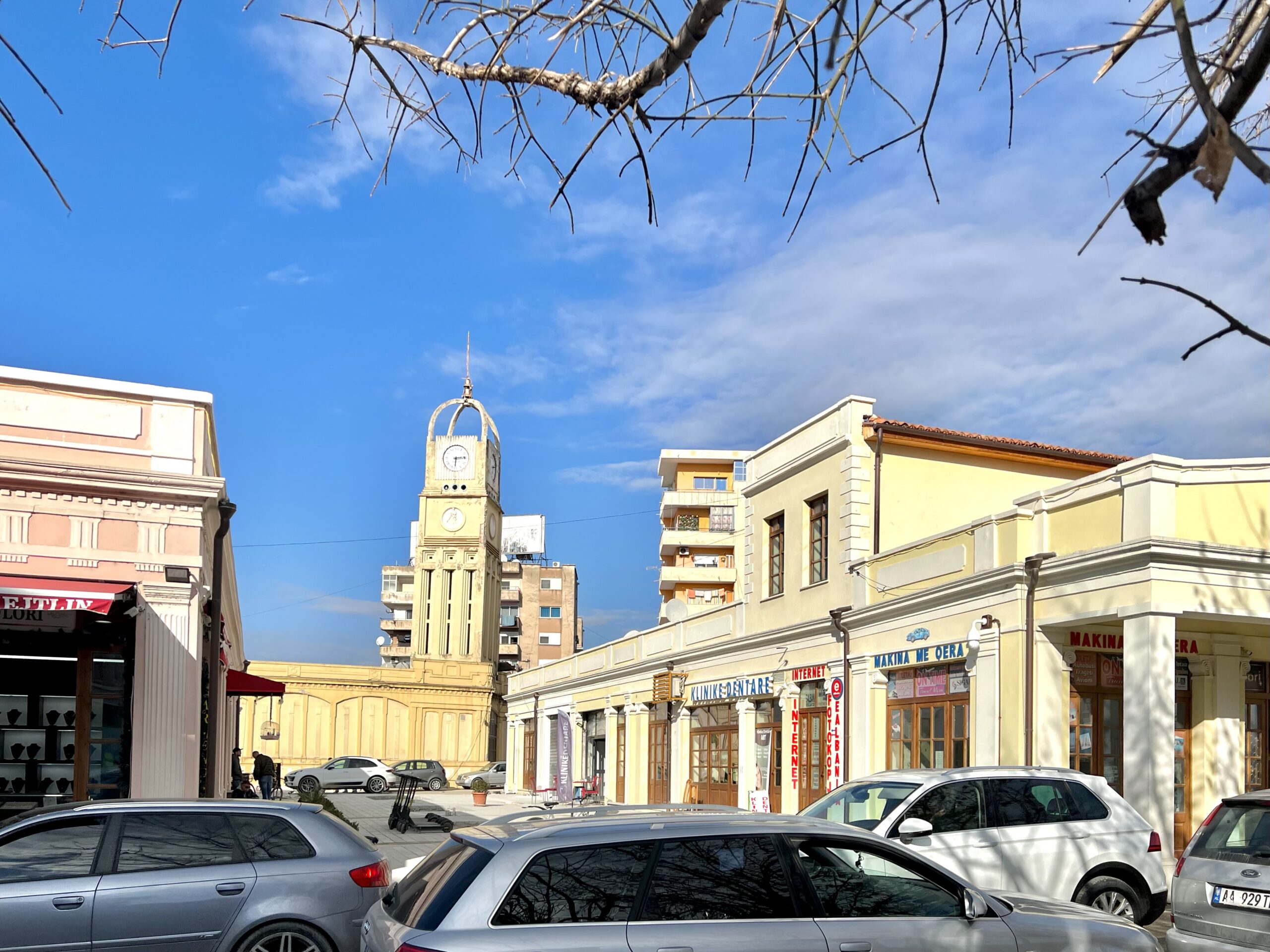 Sunny old-town square in Vlore, Albania with a clock tower, cars, and low-rise buildings.