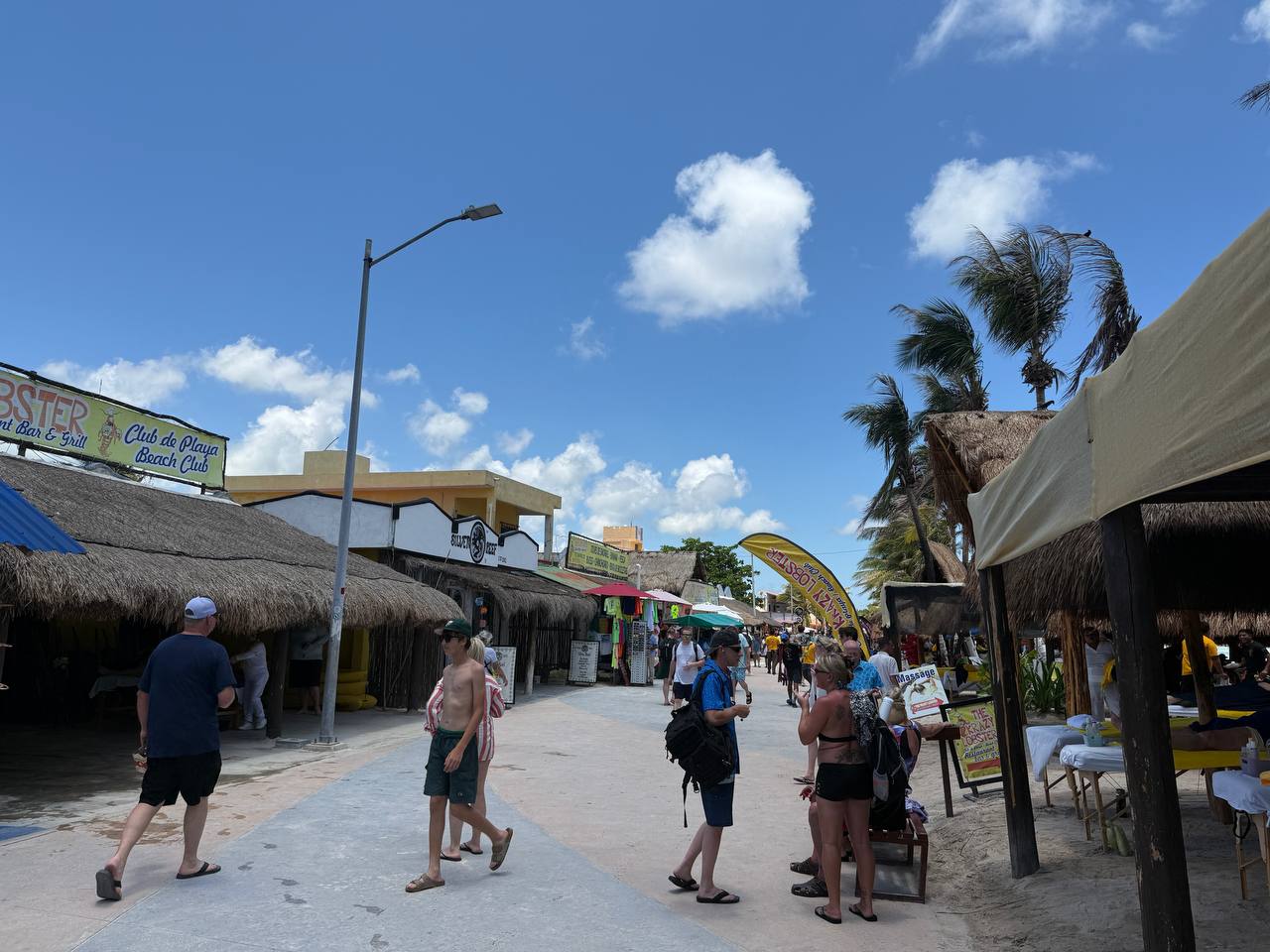 Walk-through view of the Costa Maya beach-club strip near The Krazy Lobster with palms, shops, and visitors in daylight.