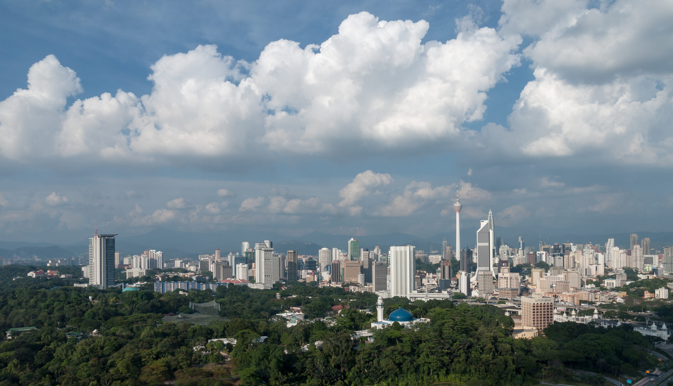 Wide skyline view of Kuala Lumpur with towers, dense urban core, and green foreground.