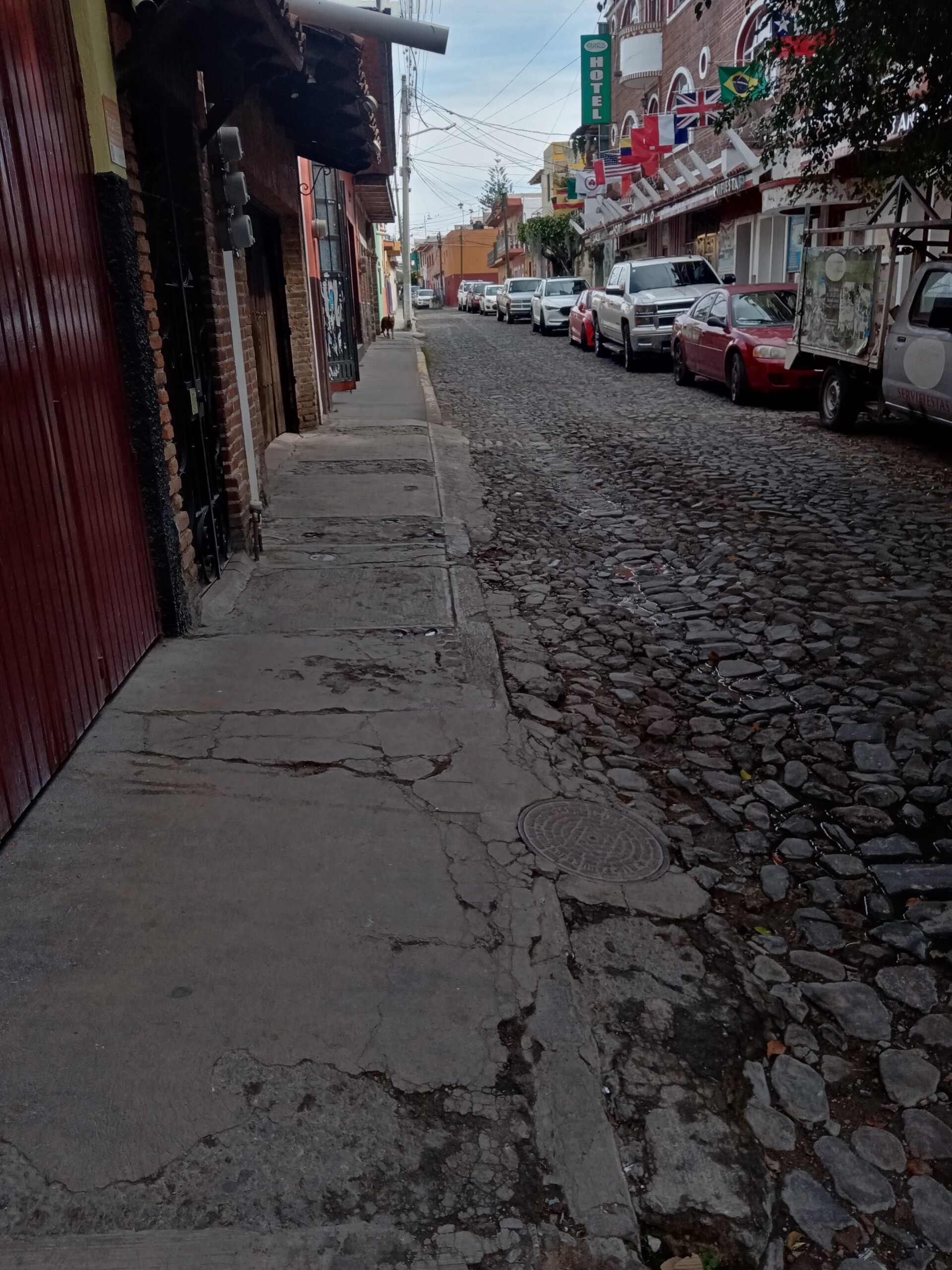 Cobblestone street and sidewalk on Calle Guadalupe Victoria in Ajijic, Jalisco, with parked cars and small-town daily-life context.