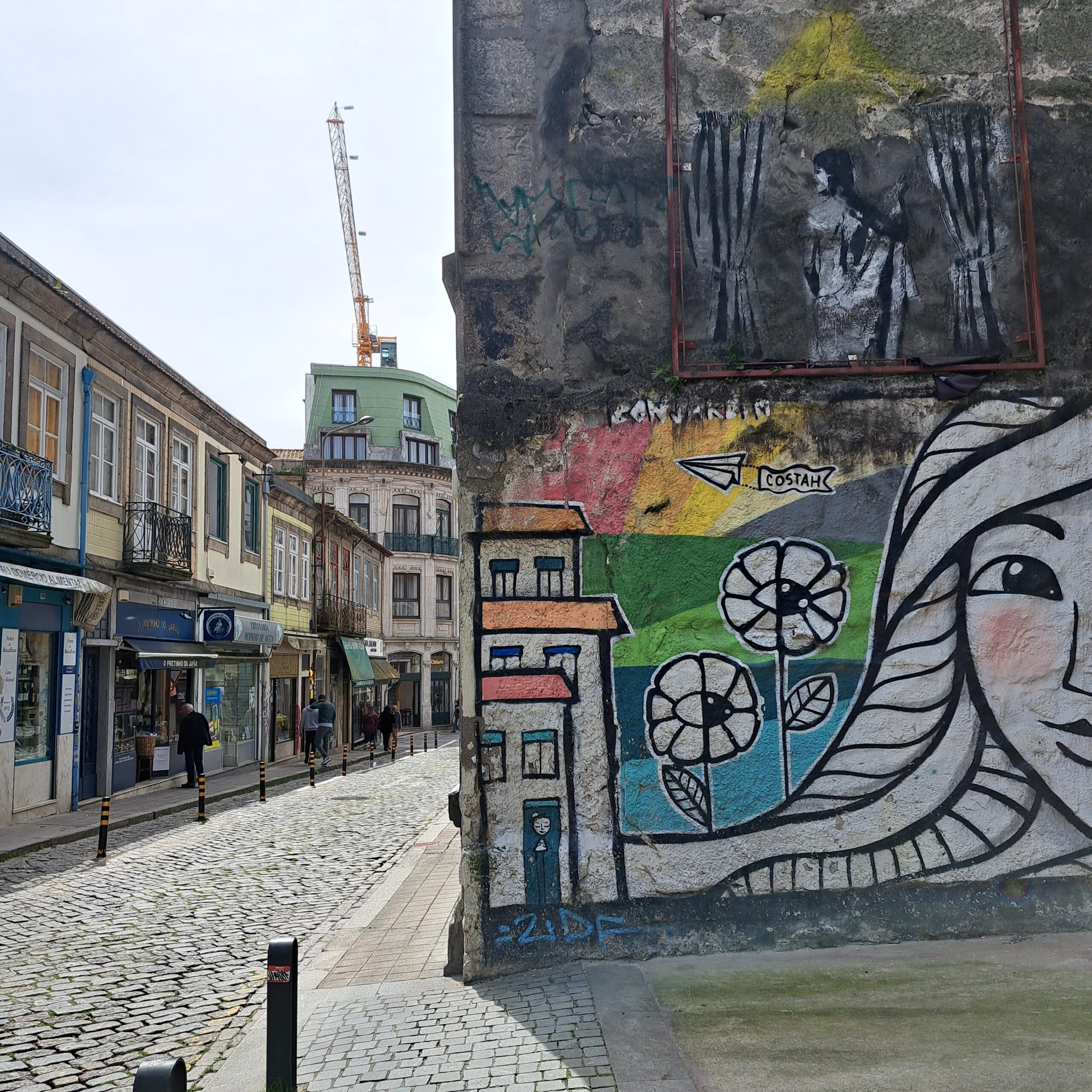 Cobblestone street in Porto with shops, pedestrians, murals, and old-city buildings.