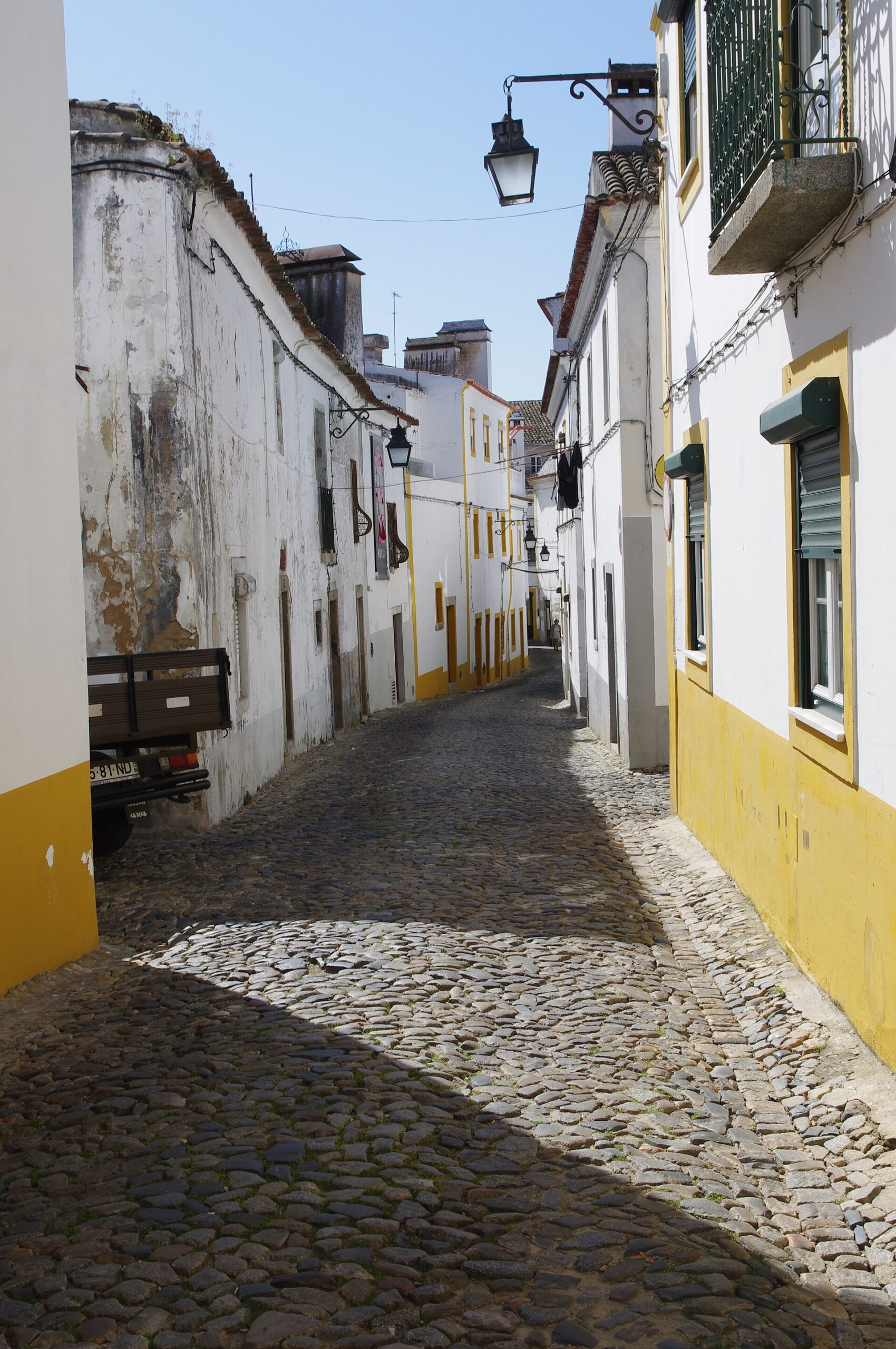 Sunlit street in Évora, Portugal with pedestrians, balconies, and a calm historic-center feel.