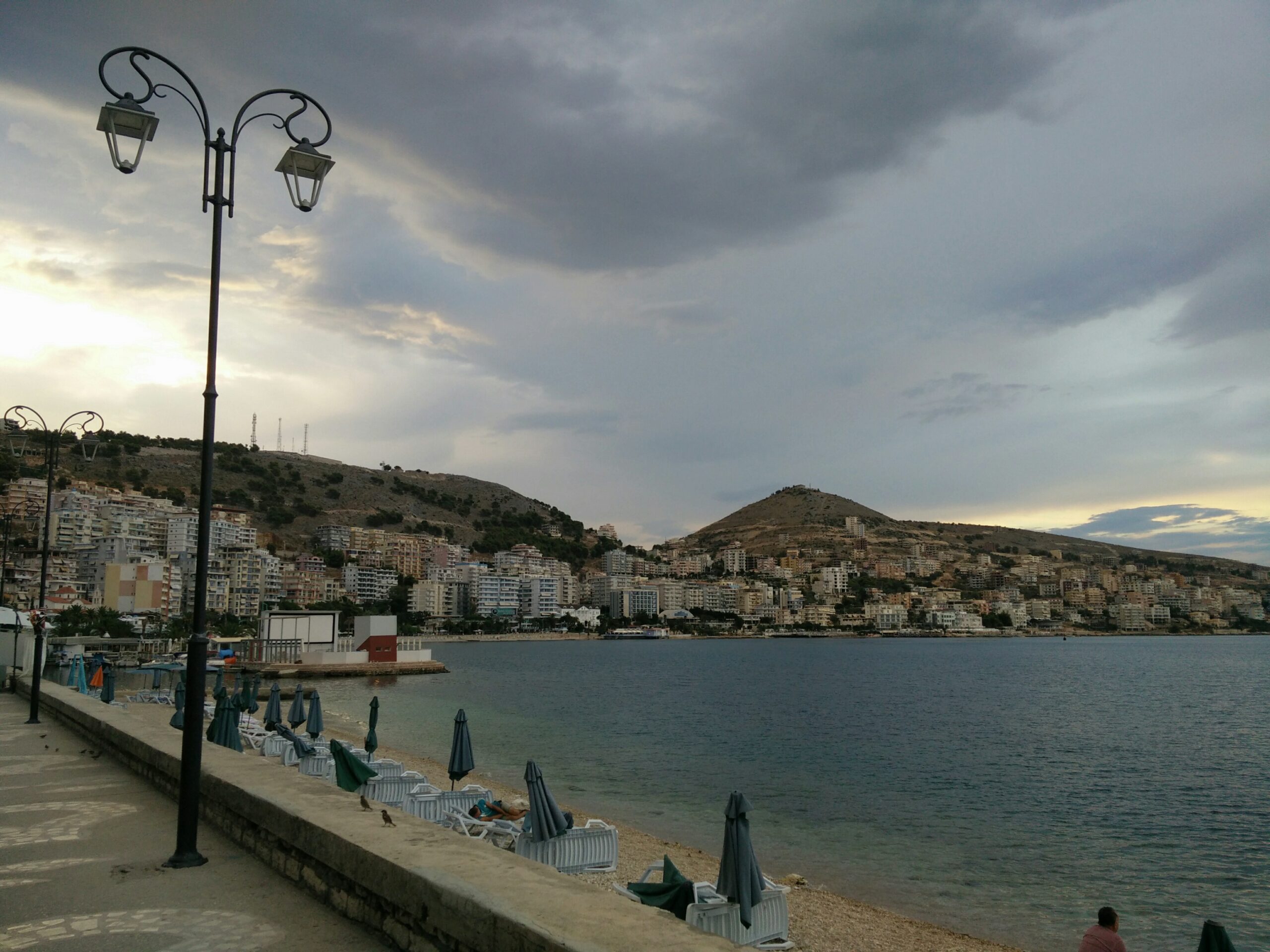 Saranda waterfront boulevard and beach with hillside buildings along the bay.