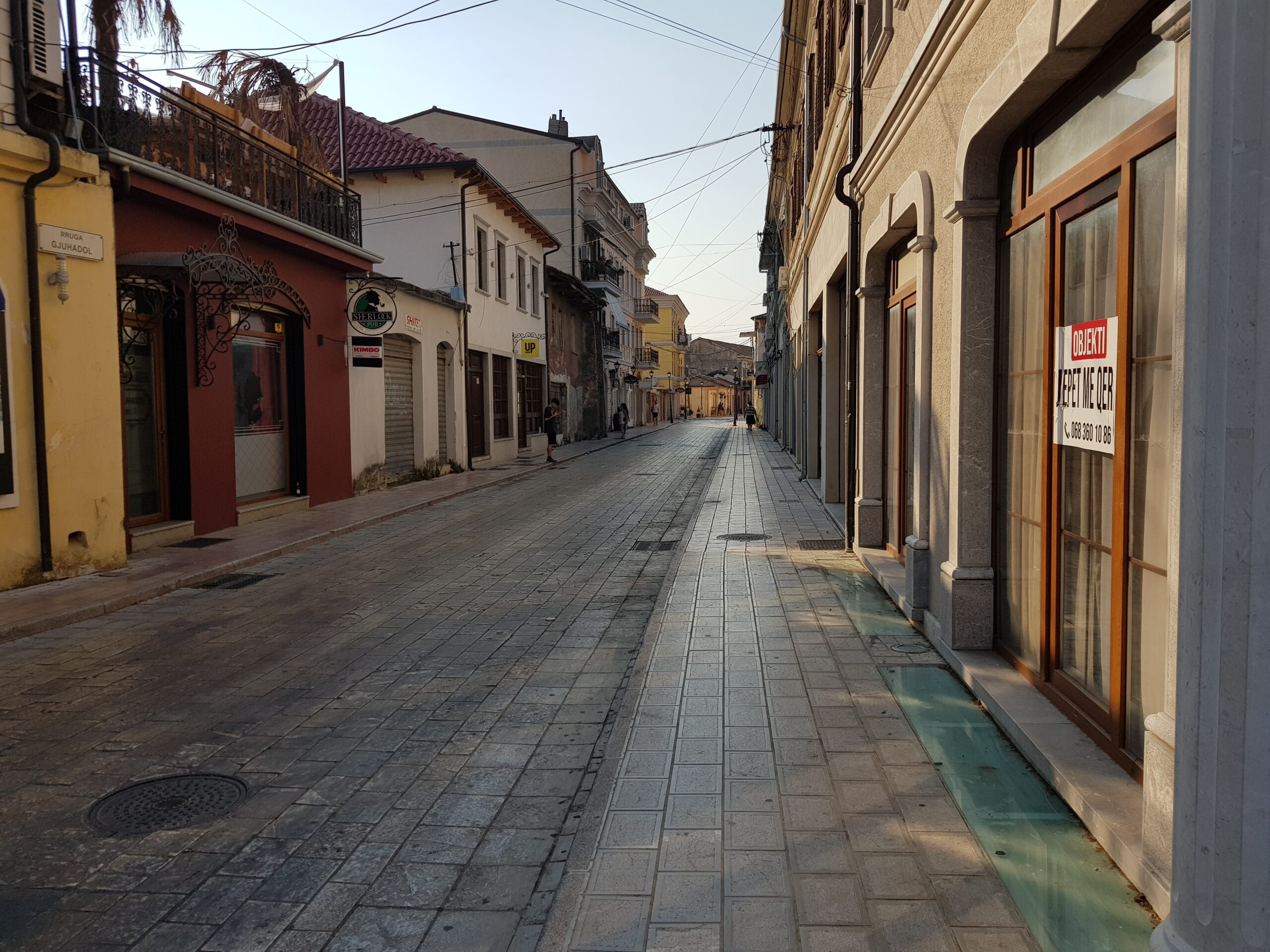 Street scene in Shkoder with storefronts, trees, and low-rise city buildings.