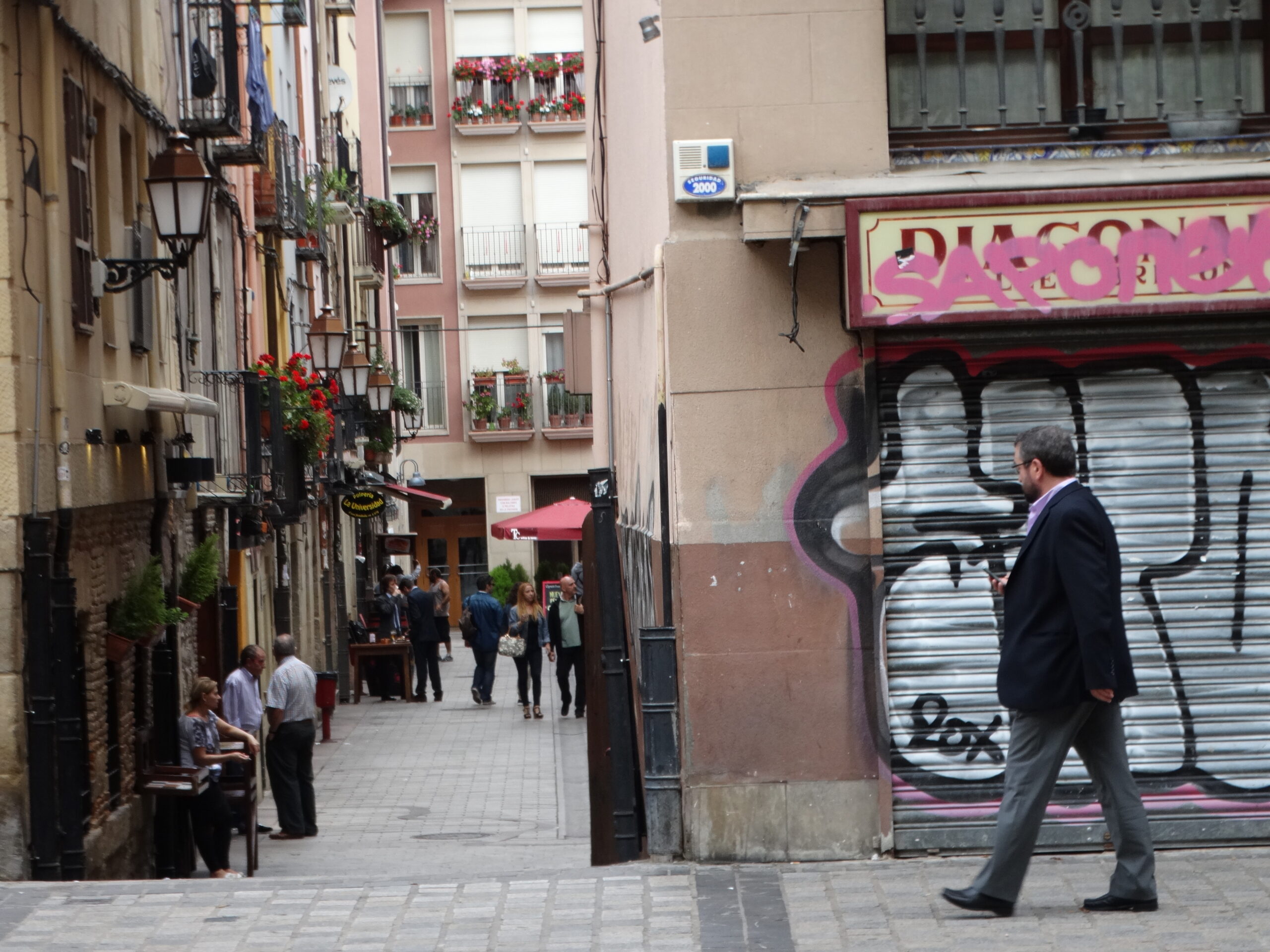 Narrow pedestrian street in Logrono, Spain with balconies, plants, storefronts, and people walking.