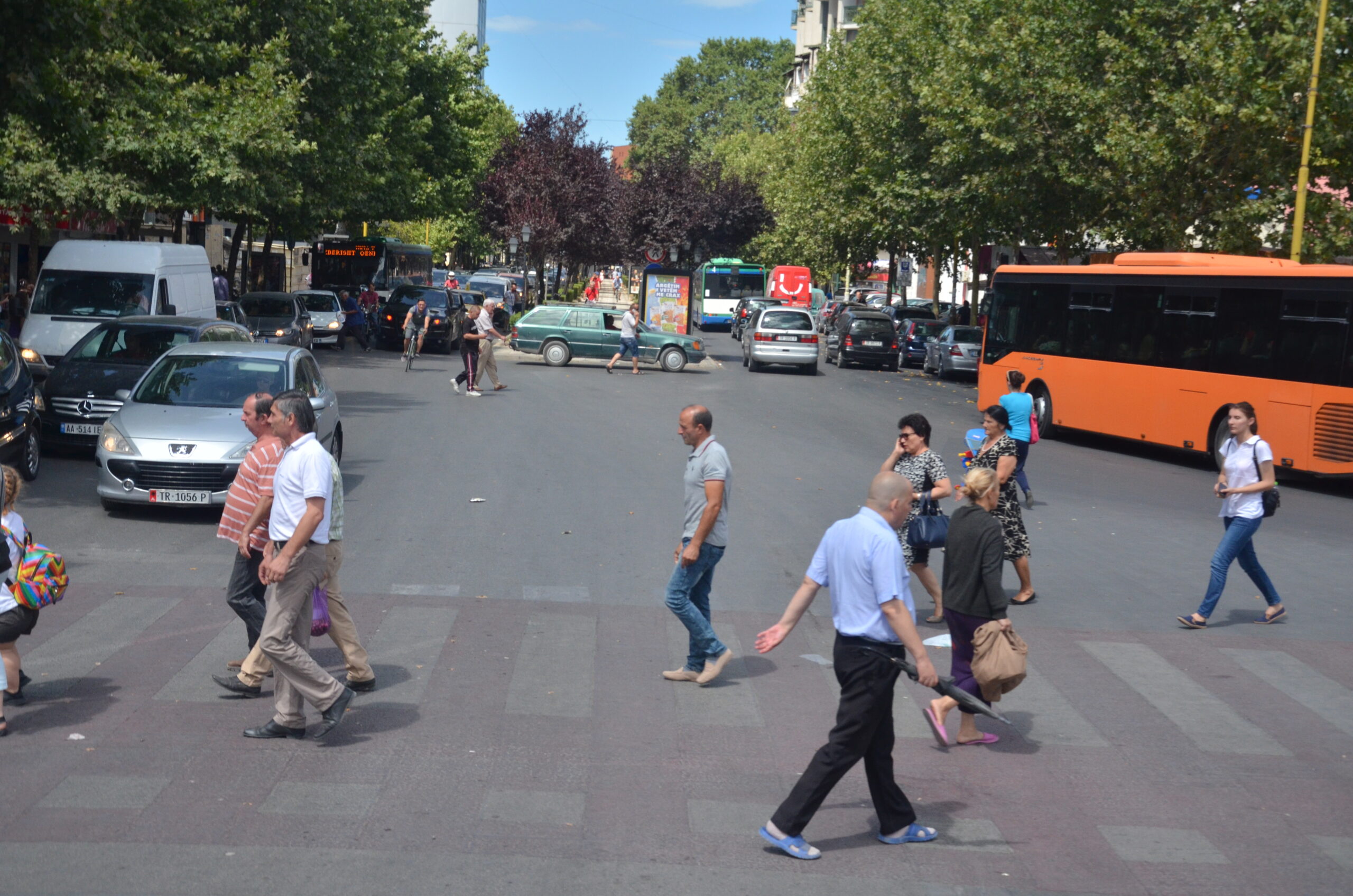 Busy Tirana street crossing with buses, pedestrians, trees, and apartment blocks.