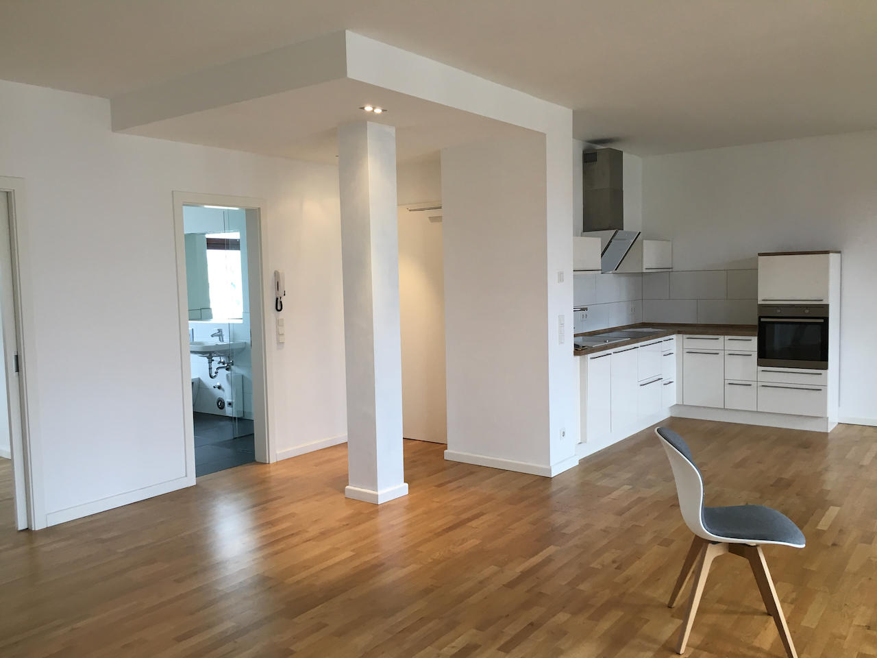 Empty apartment with fitted kitchen, bathroom doorway, open floor space, and a single chair.
