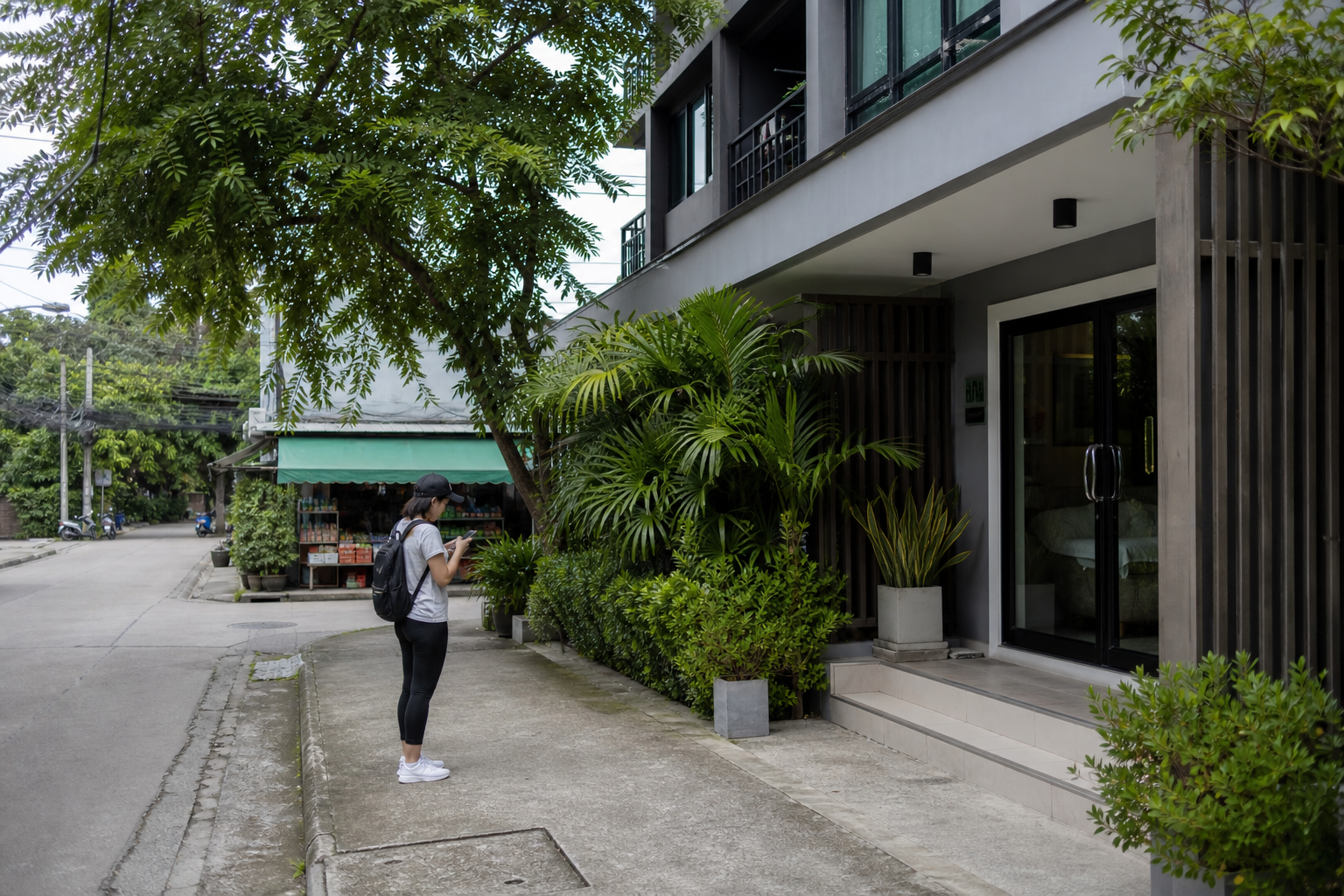 Chiang Mai apartment entrance on a leafy street, with a person checking a phone and a small grocery storefront nearby.