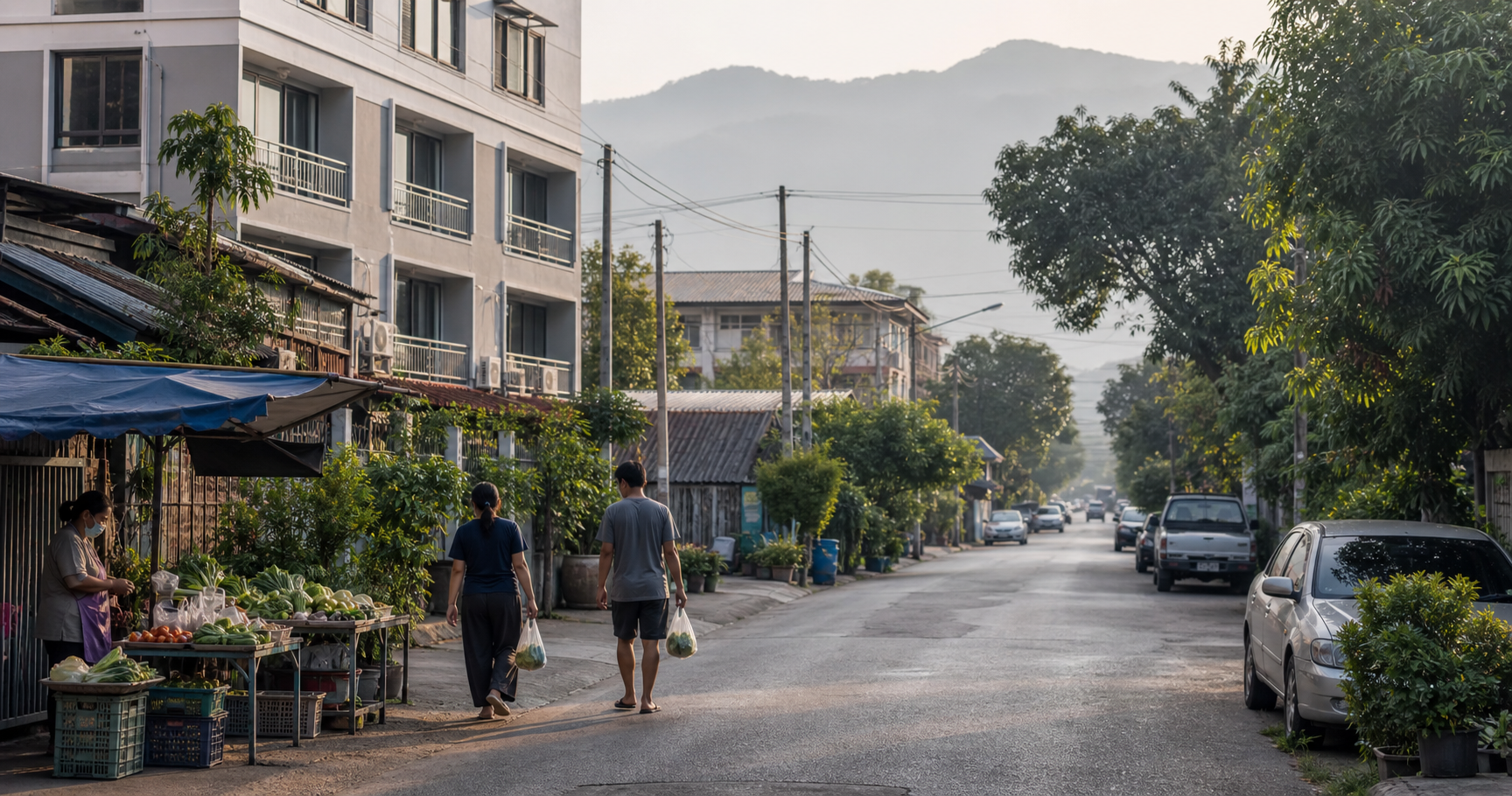 Chiang Mai neighborhood street with apartments, a produce stall, pedestrians carrying groceries, trees, and distant foothills.
