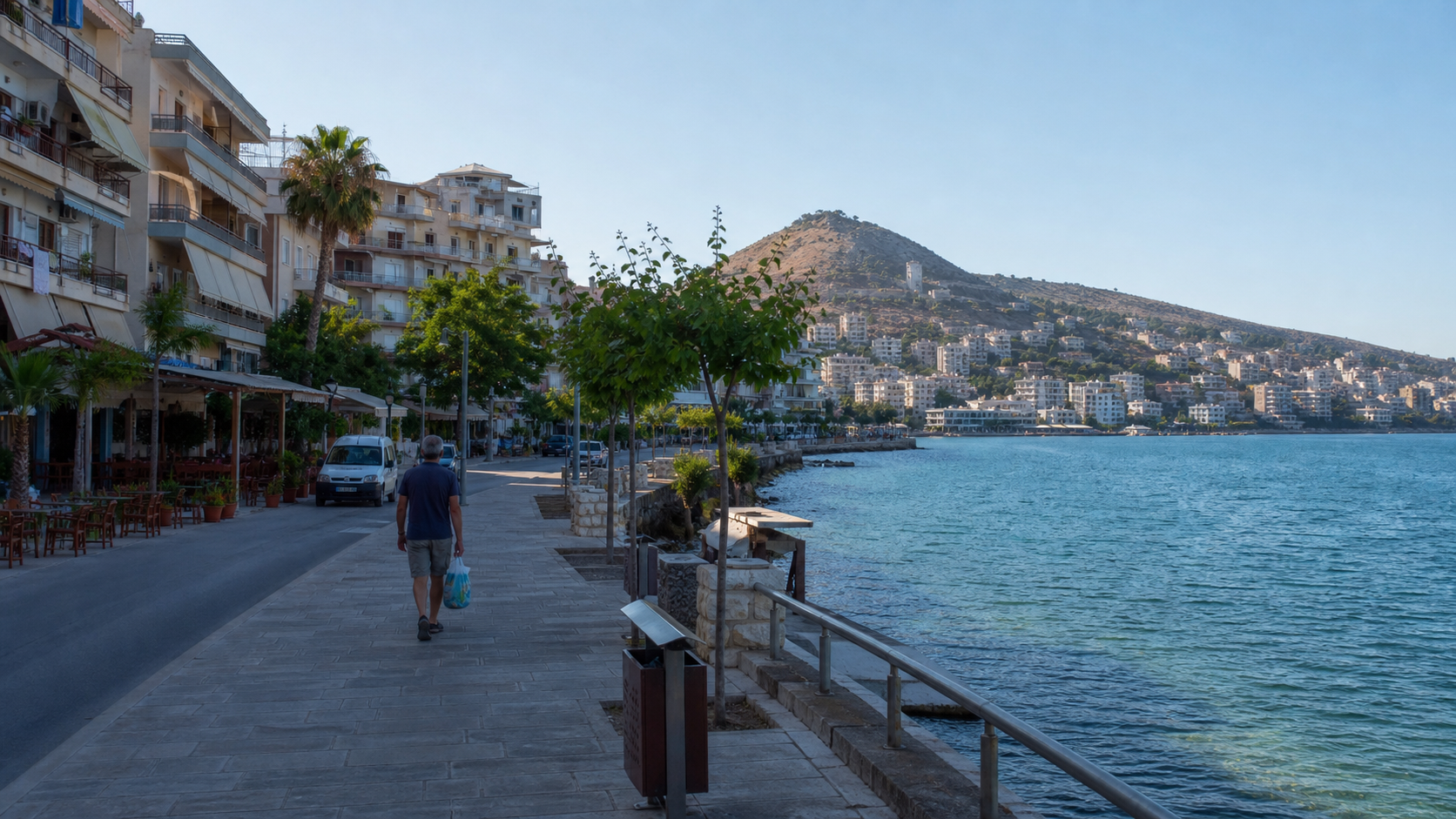 Sarande waterfront promenade with apartment buildings, a pedestrian carrying groceries, hills, and the Ionian Sea.